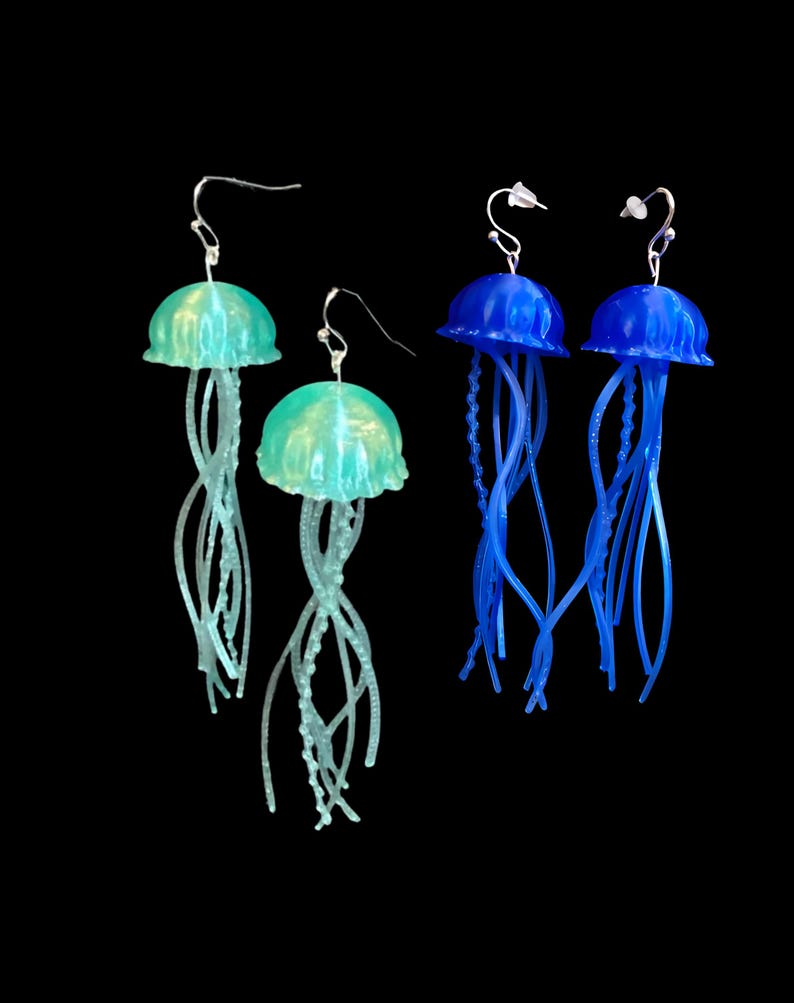 May include: A set of three dangle earrings featuring jellyfish designs. Two earrings are blue and one is green.