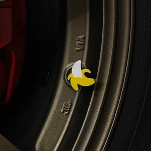 May include: A yellow banana-shaped tire valve stem cap with a white tip. The cap is attached to a black tire rim.