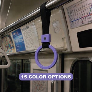 May include: A purple grab handle with a black strap, hanging from a metal bar inside a train. The handle is circular and attached to a rectangular purple base. Text at the bottom reads "15 COLOR OPTIONS".
