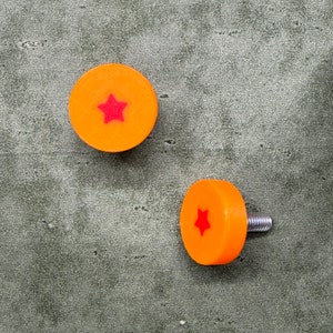 May include: Two orange cabinet knobs with a red star design. One knob is shown with a silver screw attached. The knobs are circular and have a smooth, glossy finish. The background is a mottled gray.