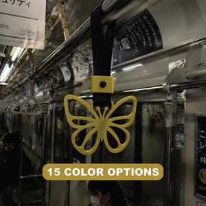 Puede incluir: Un asa amarilla con forma de mariposa para un metro o tren, colgada de una correa negra. El asa está sujeta a una barra metálica. La imagen incluye el texto "15 COLOR OPTIONS" en una pancarta amarilla.