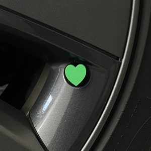 May include: A close-up of a car wheel with a green heart-shaped valve stem cap. The cap is centered on a dark gray wheel, with the tire visible on the right. The heart is a bright, solid green color.