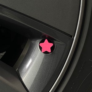May include: A bright pink star-shaped valve stem cap attached to a dark gray metallic car wheel rim. The five-pointed star features a matte finish and is positioned directly over the circular valve stem opening on the vehicle wheel.
