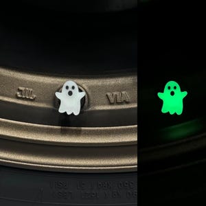 Puede incluir: Un tapón de válvula de neumático con forma de fantasma blanco brilla en verde en la oscuridad. El tapón está fijado a una válvula de neumático en una llanta de color bronce.