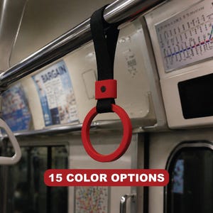Puede incluir: Un asa circular roja con una correa negra, sujeta a una barra metálica. El asa está suspendida en un vagón de metro. El texto "15 COLOR OPTIONS" se muestra en un cuadro rojo en la parte inferior de la imagen.