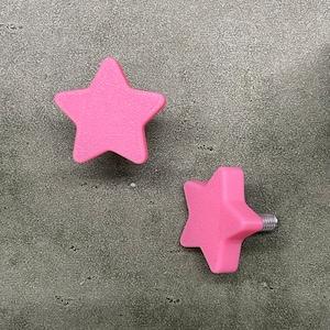 May include: Two pink star-shaped cabinet knobs. One knob is shown by itself, and the other knob has a silver screw attached. The knobs are made of a solid pink material and have a textured surface.