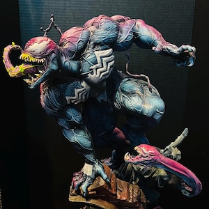 May include: A detailed, hand-painted figurine of Venom, a Marvel Comics superhero, in a dynamic pose. The figure is painted in shades of blue, purple, and black, with a textured surface to represent the character's symbiote suit. Venom is depicted leaping from a crumbling brick wall, with his mouth open and fangs bared.