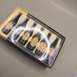 May include: A set of six miniature Guinness beer bottles in a clear plastic case with a yellow cardboard base. The bottles are black with gold labels and gold bottle caps. The labels feature the Guinness logo and other text.