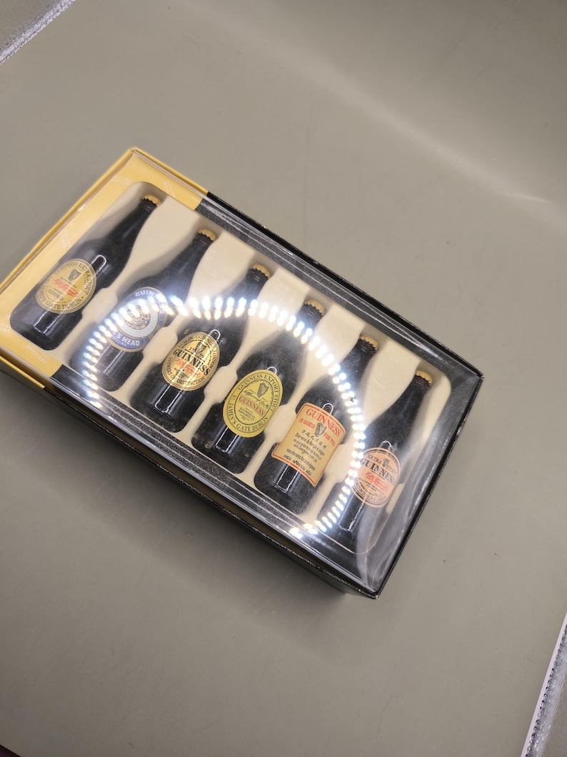 May include: A set of six miniature Guinness beer bottles in a clear plastic case with a yellow cardboard base. The bottles are black with gold labels and gold bottle caps. The labels feature the Guinness logo and other text.