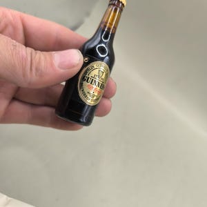 May include: Miniature Guinness beer bottle with a gold label. The label reads "Dublin Guinness 1759 Brewers of Distinction". The bottle is dark brown with a gold cap and a clear neck.