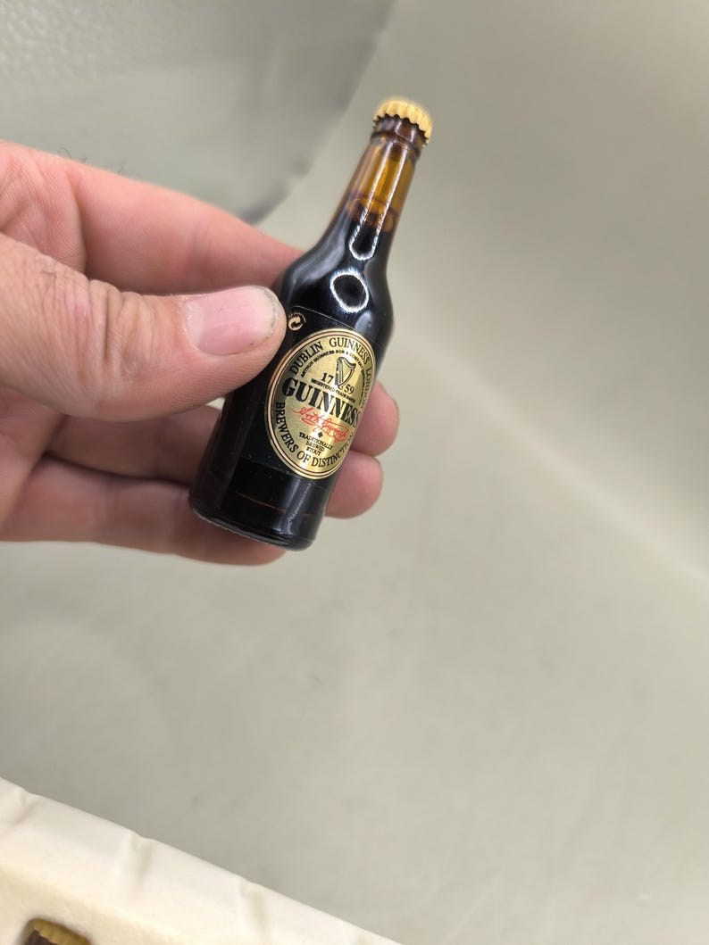 May include: Miniature Guinness beer bottle with a gold label. The label reads "Dublin Guinness 1759 Brewers of Distinction". The bottle is dark brown with a gold cap and a clear neck.