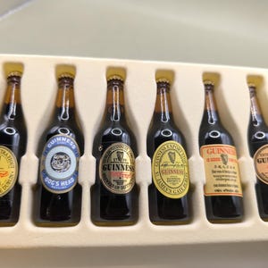 May include: A set of six miniature Guinness stout bottles in a cream-colored tray. Each bottle has a different label, including "Extra Stout," "Dog's Head," and "Guinness Export." The labels feature gold, yellow, and blue accents.