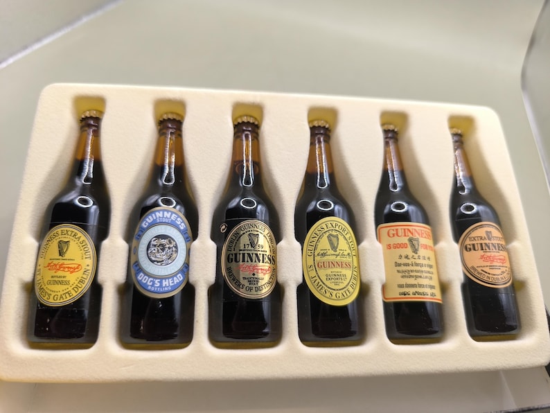 May include: A set of six miniature Guinness stout bottles in a cream-colored tray. Each bottle has a different label, including "Extra Stout," "Dog's Head," and "Guinness Export." The labels feature gold, yellow, and blue accents.