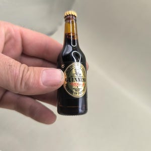May include: A miniature Guinness beer bottle with a gold cap and a black and gold label. The label reads "Guinness" and "Brewery of Distinction". The bottle is filled with a dark liquid.