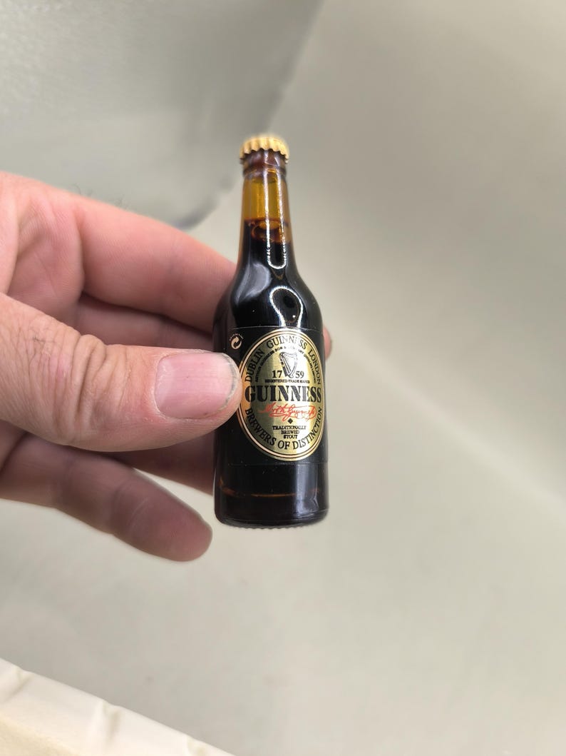 May include: A miniature Guinness beer bottle with a gold cap and a black and gold label. The label reads "Guinness" and "Brewery of Distinction". The bottle is filled with a dark liquid.