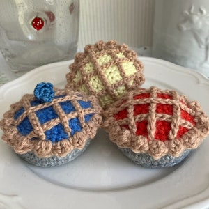 May include: Three crocheted mini pies on a white plate. The pies are in the colors red, blue, and yellow. The pies have a lattice crust and are decorated with a small blue crocheted blueberry on top of the blue pie.