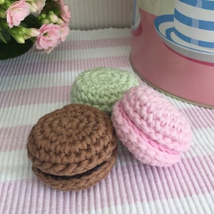 May include: Three crocheted macarons in pink, green, and brown. The macarons are sitting on a pink and white striped surface.