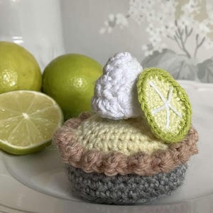 May include: Crocheted lime pie with a white crocheted lime wedge on top. The pie is sitting on a white plate with limes in the background.