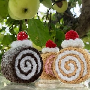 Könnte beinhalten: Drei gehäkelte Kuchenrollen-Anhänger mit Erdbeer- und Schlagsahne-Topping. Die Kuchenrollen sind braun, rosa und beige.