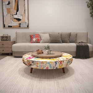 Puede incluir: Escena de salón con una mesa de centro redonda con una parte superior estampada colorida y patas de madera. Un sofá gris claro con cojines y una manta se encuentra detrás de la mesa. También se ve una mesa auxiliar de madera y arte abstracto.