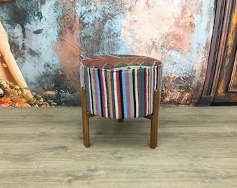 May include: A round, colorful, striped ottoman with a wooden base. The ottoman is made of woven fabric with a variety of colors, including pink, blue, green, and brown. The wooden base is a dark brown color.