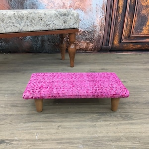 May include: A small pink footstool with wooden legs. The footstool is covered in a patterned fabric.