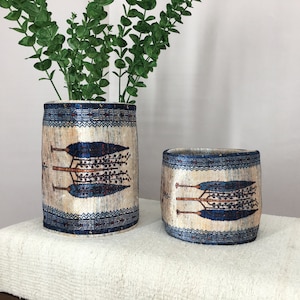 Könnte beinhalten: Zwei dekorative zylindrische Vasen mit einem blauen und weißen Muster. Das Muster zeigt stilisierte Bäume mit blauen Blättern und braunen Ästen. Die Vasen stehen auf einer weißen Oberfläche.