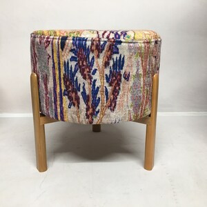 May include: A round, upholstered ottoman with a colorful floral pattern in shades of blue, red, and yellow. The ottoman has four wooden legs.