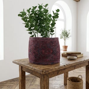 May include: A burgundy felt planter pot with a dark pattern holds a lush green plant. The pot sits on a rustic wooden table. The room has a bright, airy feel with a window in the background.