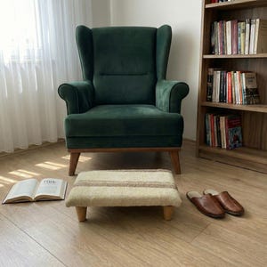 May include: A deep green armchair with wooden legs and a coordinating footstool. The footstool features a beige and brown striped design. Brown leather slippers and an open book are on the floor. A wooden bookshelf is in the background.