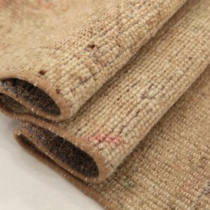 Op de afbeelding: Een gevouwen geweven tapijt met een getextureerde pool in tinten beige, zand en lichtbruin. De randen van het tapijt hebben een donkerbruine afwerking en het oppervlak vertoont subtiele hints van vervaagde roze en gedempte groene patronen.
