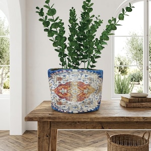 May include: A decorative, cylindrical plant pot with a blue rim and a patterned design in red, yellow, and white. The pot is filled with green leafy plants and sits on a wooden table. A wicker basket is in the foreground.