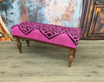 May include: A pink and black patterned ottoman with wooden legs. The ottoman is made of a soft, woven fabric and has a geometric design.