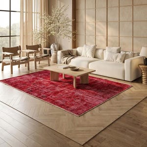 Może przedstawiać: Salon z dużym, czerwonym dywanem. Dywan jest umieszczony przed kremową sofą i drewnianym stolikiem kawowym. Widoczne są dwa drewniane krzesła oraz duży wazon z białymi kwiatami.