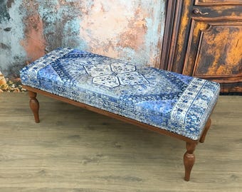 May include: A rectangular bench featuring a cushioned seat upholstered in a blue and white fabric with an intricate, symmetrical medallion pattern. The bench is supported by four turned wooden legs with a warm, medium-brown finish.