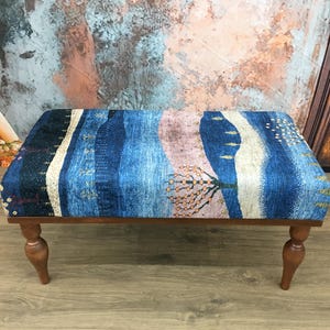 Puede incluir: Un reposapi&eacute;s de madera con patas torneadas y una parte superior de tela con estampado azul y rosa. La tela presenta un dise&ntilde;o de paisaje con &aacute;rboles y colinas.