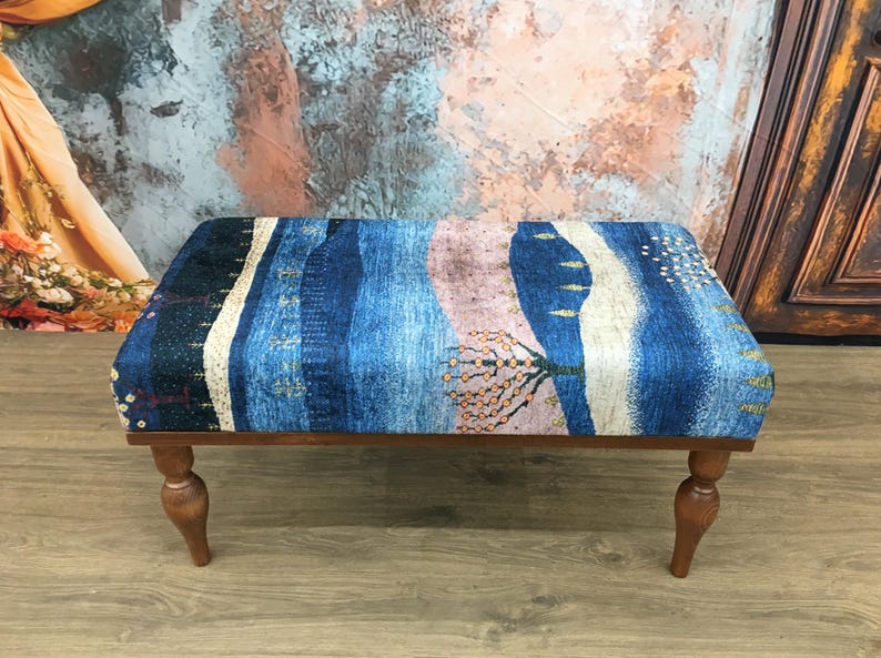 Puede incluir: Un reposapi&eacute;s de madera con patas torneadas y una parte superior de tela con estampado azul y rosa. La tela presenta un dise&ntilde;o de paisaje con &aacute;rboles y colinas.