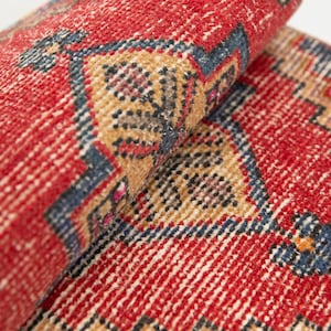 May include: A woven rug featuring a vibrant red background with intricate geometric patterns in shades of navy blue, beige, and hints of orange. The rug is partially rolled, revealing a textured pile surface with a repeating floral-inspired motif.