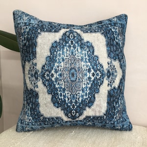 Op de afbeelding: Vierkant decoratief kussen met een blauw en wit patroon. Het kussen heeft een centraal medaillonontwerp met bloem- en geometrische elementen. Het kleurenpalet omvat tinten blauw, wit en grijs, wat een vintage esthetiek cre&euml;ert.