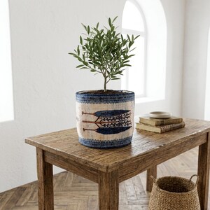 May include: A small olive tree in a decorative pot with a blue and cream pattern. The pot sits on a wooden table with a woven basket and books. The pot has a blue patterned rim and a design of blue feathers.