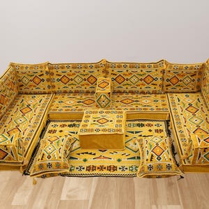 Puede incluir: Un juego de sofás modular amarillo con intrincados patrones geométricos en naranja, verde y blanco. El conjunto incluye cojines, una otomana central y una alfombra estampada, creando una disposición de asientos cohesiva. Adecuado para una sala de estar o salón.