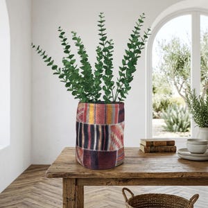 May include: A decorative vase with a patchwork design in shades of red, yellow, and black, holding green eucalyptus branches. The vase sits on a wooden table, creating a rustic and natural aesthetic.