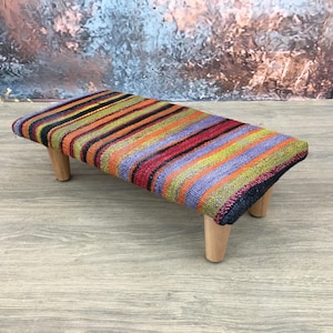 May include: A small, rectangular footstool with a colourful striped pattern in shades of red, orange, green, and purple. The stool has four short, light-coloured wooden legs. The design combines rustic and vibrant elements.