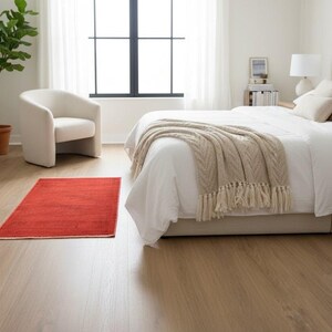 May include: A bedroom interior featuring light brown wood-look flooring with a rectangular red area rug. The room includes a bed with white bedding and a beige cable-knit throw blanket, a cream-colored armchair, and a potted green plant.