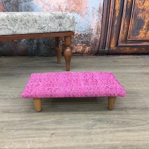 May include: A pink upholstered footstool with wooden legs. The footstool is sitting on a wooden floor.