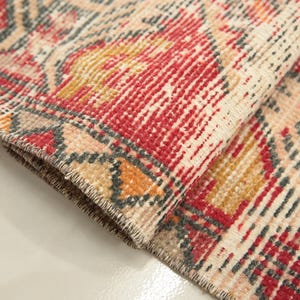 May include: A woven rug featuring a detailed pattern in shades of red, mustard yellow, and charcoal gray on a cream background. The rug is folded over to reveal the textured pile and the finished edge with visible stitching along the border.