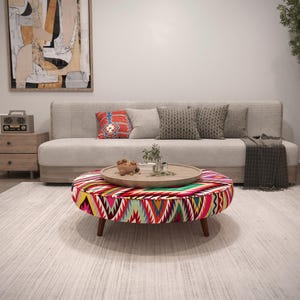 May include: A living room scene featuring a beige sofa with decorative pillows and a patterned ottoman. The ottoman has a colorful geometric design. A wooden tray with plants and decor sits on top. A large abstract painting hangs on the wall.