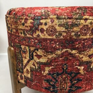 May include: A round ottoman with a red, gold and blue patterned fabric cover. The ottoman has a wooden base with dark brown legs.