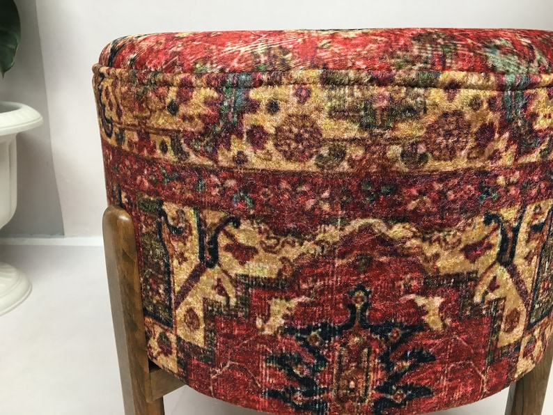 May include: A round ottoman with a red, gold and blue patterned fabric cover. The ottoman has a wooden base with dark brown legs.