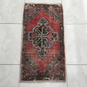 May include: A small red rug with a geometric pattern in shades of black, blue, and purple. The rug has a fringe border.
